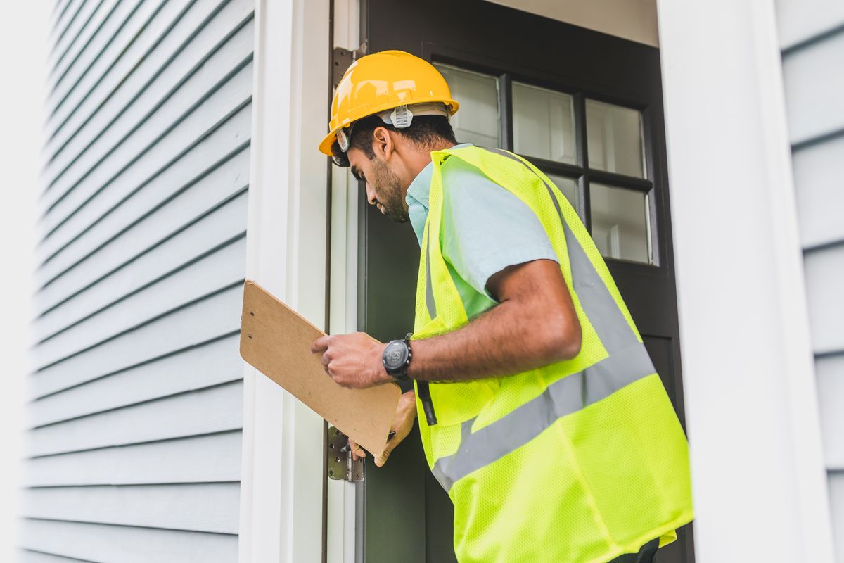 Inspector wearing a yellow hard hat and reflective vest looking closely at an exterior doorway while holding a clipboard