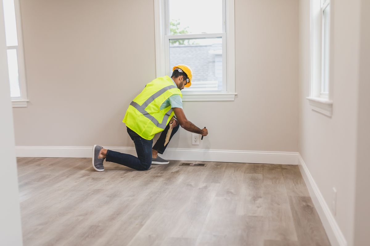 Inspector in a safety vest kneeling by a wall in an empty room, checking an electrical outlet near a floor vent